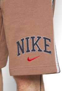 Braune Sportshorts mit gesticktem "NIKE" in Dunkelblau und kleinem roten Swoosh-Logo, weißen Seitenstreifen und strukturiertem Finish.