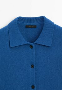 Cardigan bleu en maille avec col plié et trois boutons noirs sur le devant, affichant l'étiquette de la marque à l'intérieur de l'encolure.