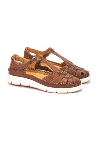 Sandalias de cuero marrón con diseño recortado, acentos trenzados y correa ajustable. Suela gruesa blanca con una base marrón texturizada.