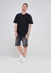 Juodas per didelis marškinėliai trumpomis rankovėmis, derinami su tamsiai pilkais džinsiniais šortais. Balti sportbačiai puikiai papildo šį įvaizdį. Lygus fonas.