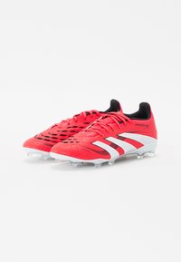 Chaussures de football rouges avec un dessus texturé, des accents noirs et des rayures blanches. Comprend un design à lacets et des crampons en plastique transparent.