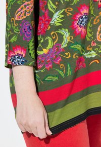 Kleurige bloemenprint top in groen met rode en groene horizontale strepen aan de zoom. Beschikt over driekwartmouwen en een zachte textuur.
