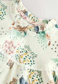 Kindermode in cremefarbenem Stoff mit floralem Muster, das Tiere, Bäume und einen Rüschenausschnitt zeigt, der mit Knopfverschlüssen am Hals befestigt ist.