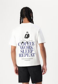 Weißes Baumwoll-T-Shirt mit einem Graf design, das eine sitzende Figur und den Text "KAFFEE ARBEITEN SCHLAFEN WIEDERHOLEN" in Blau zeigt. Kurze Ärmel, lockere Passform.