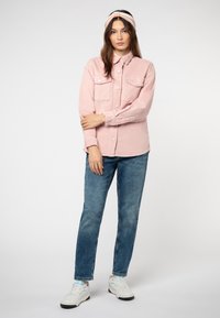Lichtroze corduroy overhemd met knoopsluiting aan de voorkant, twee borstzakken en lange mouwen. Gecombineerd met blauwe denim jeans en witte sneakers.