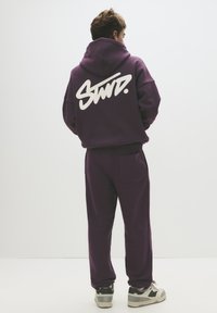 Hanorac mov și pantaloni sport asortați, confecționați dintr-un material moale, având un logo alb mare pe spate. Adidași gri completează ținuta.