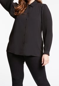 Svart blus med knäppning fram och klassisk krage. Har texturerade plisserade långärmar och slätt kroppstyg.