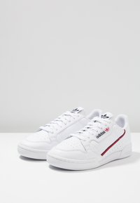 White sneakers with smooth leather upper, perforated toe box, contrasting red and navy stripe, cushioned collar, and flat rubber sole.