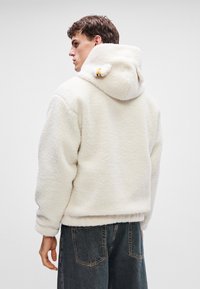 Hanorac pufos alb cu buzunar tip cangur, glugă ajustabilă cu urechi mici și o croială lejeră, asortat cu blugi din denim închisi la culoare.