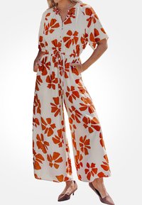 Witte jumpsuit met grote oranje bloemprint, korte mouwen, elastische taille en zijzakken. Gecombineerd met bruine blockhakken.