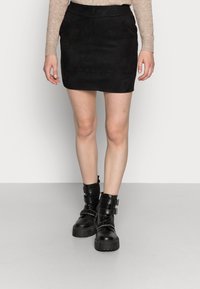 Jupe mini en daim noir au design ajusté. Associée à des bottes noires volumineuses avec des boucles et des semelles texturées.