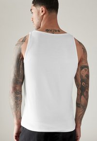 Weiße ärmellose Tanktop aus glattem Material mit einem schlichten Design. Die tätowierten Arme eines Mannes sind sichtbar, mit komplexen Mustern auf der Haut.