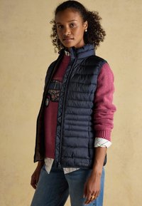 Young woman wearing a navy quilted vest over a pink sweater with a car design, paired with blue jeans and a checked shirt underneath.