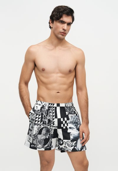 Jeune homme torse nu portant un short noir et blanc à motifs avec des visages artistiques et des dessins géométriques, debout devant un fond uni.
