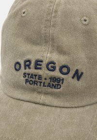 Beige színű pamut baseball sapka, tengerészkék hímzett felirattal: "OREGON STATE 1991 PORTLAND." A felirat a sapka elülső paneljén található.