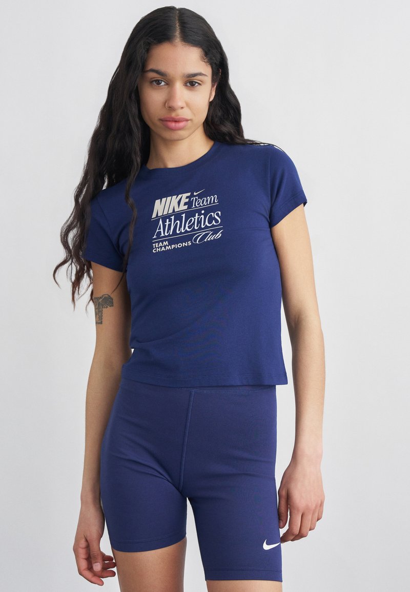 Γυναίκα με μακριά σκούρα μαλλιά φοράει μπλε σκούρο T-shirt Nike Athletics και αντιστοιχισμένο σορτς ποδηλασίας, στέκεται μπροστά σε απλό φωτεινό φόντο.