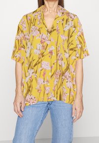 Kurzarmshirt aus gelbem Stoff mit floralem Muster und pinken Schwertlilien, mit Knopfleiste und lässiger Passform. Kombiniert mit blauen Jeans.