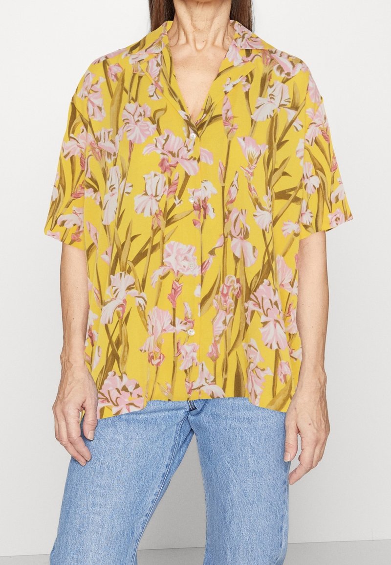 Kurzarmshirt aus gelbem Stoff mit floralem Muster und pinken Schwertlilien, mit Knopfleiste und lässiger Passform. Kombiniert mit blauen Jeans.