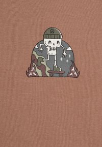 Καφέ μπλούζα T-shirt με γραφικό ενός καρτούν σκελετού που κάνει σκέιτ, φορώντας πράσινο καπέλο, μπροστά σε στρογγυλό φόντο που περιλαμβάνει φλόγες και σύννεφα.