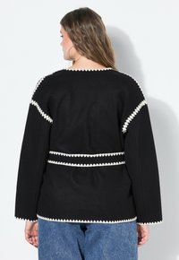 Femme avec de longs cheveux bruns portant une veste noire avec un liseré blanc en zigzag, se tenant contre un mur blanc, tournant le dos à la caméra.