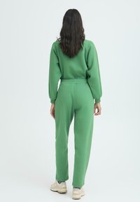 Zeleni trenirku s dugim rukavima, elastičnim strukom i suženim nogavicama, izrađenu od mekog, teksturiranog materijala. Vidljive šavove.