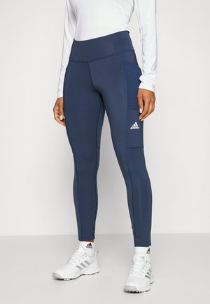 Woman wearing navy high-waist Adidas leggings with side pocket, white long-sleeve top, white Adidas socks, and white athletic shoes.