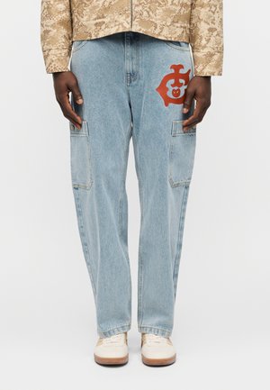 Lyseblå denim cargobukser med sidelommer og et rødt emblem på øverste lår, båret med beige mønstret jakke og hvide sneakers.