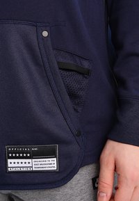 Marinblå sweatshirt med framficka, nätdekoration och en patchetikett. Har en slät yta och kontrasterande detaljer.