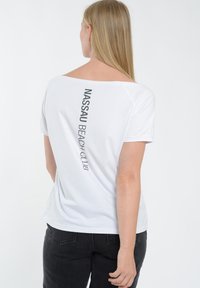 Hvid T-shirt med bred halsudskæring og korte ærmer, der har vertikal sort tekst "NASSAU BEACH CLUB" på ryggen. Blødt bomuldsstof.