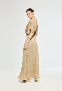 Beige satin dress with dolman sleeves, flowing silhouette, and back cutout detail. Model wearing gold accessories and strappy sandals.