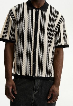 Mann trägt ein kurzärmeliges gestricktes Hemd mit schwarzen und weißen vertikalen Streifen, schwarzem Kragen und Knopfleiste, kombiniert mit einer dunklen Hose.