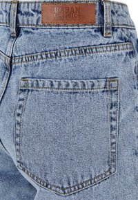 Světle modré džíny z denimu s obdélníkovou koženou etiketou s nápisem "URBAN CLASSICS". Na zadní kapse je viditelný šev.