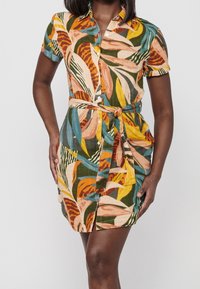 Femme portant une robe à manches courtes, boutonnée, avec un motif coloré de feuilles et une ceinture en tissu nouée à la taille.