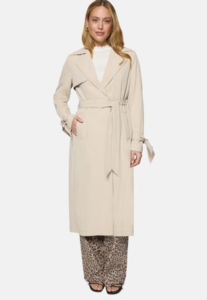 Femme portant un trench-coat beige ceinturé, un haut blanc, un pantalon à jambes larges imprimé léopard et des talons ouverts noirs, debout devant un fond uni.