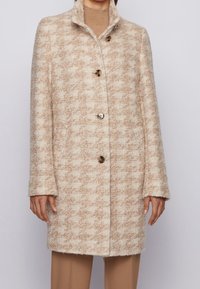 Beige og off-white tekstureret uldfrakke med høj krave og fire store knapper iført over en tan rullekrave og bukser.