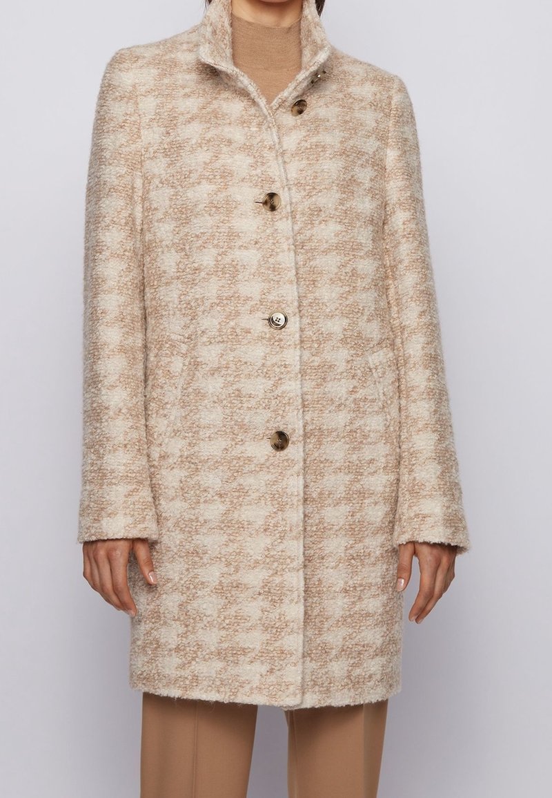 Beige og off-white tekstureret uldfrakke med høj krave og fire store knapper iført over en tan rullekrave og bukser.