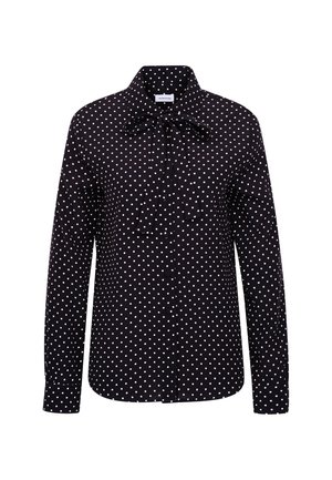 Donna in piedi che indossa una camicetta nera a pois a maniche lunghe con fiocco, pantaloni neri su misura a vita alta e mocassini neri su sfondo bianco.