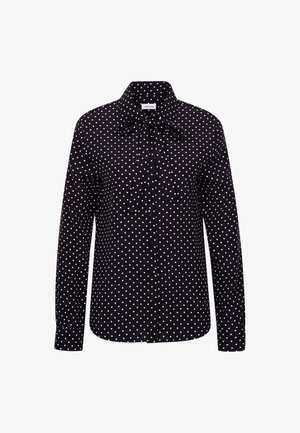 Kvinde står iført sort polkaprikket bluse med lange ærmer og sløjfe, sorte højtaljede skræddersyede bukser og sorte loafers mod hvid baggrund.