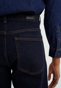 Tmavé džíny z denimu s oranžovým šitím, zadní kapsou s zakřiveným klopou a koženou nášivkou na pasu.
