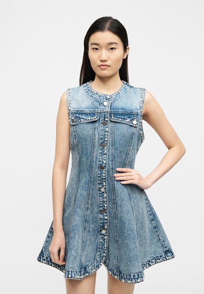 Νεαρή γυναίκα με μακριά σκούρα μαλλιά που φοράει αμάνικο μπλε φόρεμα από τζιν με διακοσμητικά μαργαριταρένια στοιχεία και κουμπιά μπροστά, στέκεται με το ένα χέρι στη μέση.