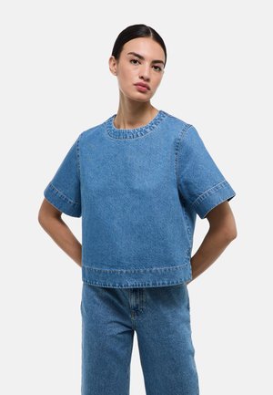 Femme aux cheveux foncés attachés en chignon bas, portant une chemise en jean bleu à manches courtes et un pantalon en jean bleu assorti, debout les mains derrière le dos.