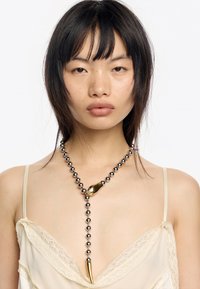 Srebrny naszyjnik z kulkowym łańcuchem z złotym akcentem w abstrakcyjnym kształcie oraz zwężającym się wisiorkiem. Modelka nosi jasną bluzkę z koronkowym wykończeniem.