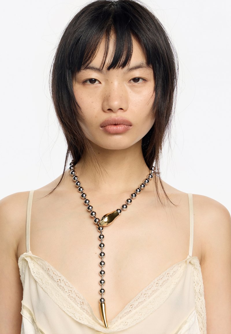 Srebrny naszyjnik z kulkowym łańcuchem z złotym akcentem w abstrakcyjnym kształcie oraz zwężającym się wisiorkiem. Modelka nosi jasną bluzkę z koronkowym wykończeniem.