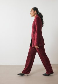 Traje de terciopelo carmesí con un acabado texturizado, que incluye una chaqueta entallada y pantalones de pierna ancha combinados con tacones negros de punta.