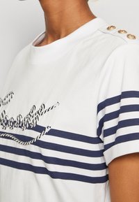 Bílá tričko s námořnickými pruhy, s texturovaným designem lana a akcenty zlatých knoflíků na rameni. Krátké rukávy a kulatý výstřih.