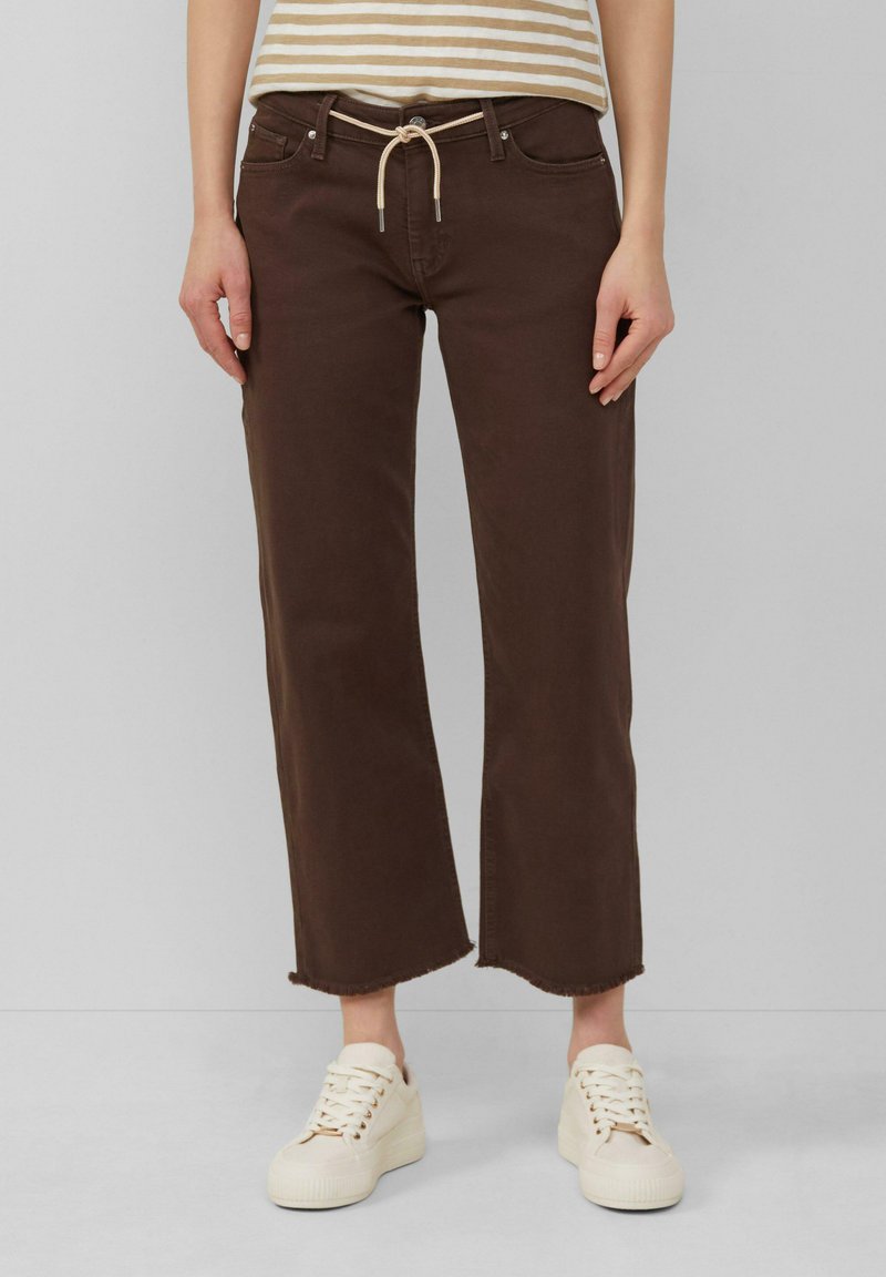 Brązowe spodnie szerokie w nogawkach z elastycznym paskiem i strzępionym dołem, w zestawieniu z białymi trampkami. Wyglądają na gładkie i przypominające bawełnę.