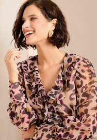 Priehľadná blúzka s ružovo-čiernym leopardím vzorom, volánovým výstrihom a našitými dlhými rukávmi s gombíkovými detailmi. Zlaté náušnice dodávajú štýl.