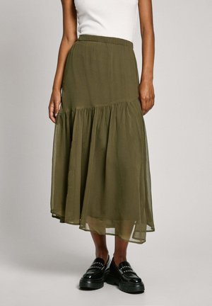 Femme portant une jupe midi à volants vert olive, un haut sans manches blanc et des mocassins plateformes noirs cloutés, debout sur un fond gris.