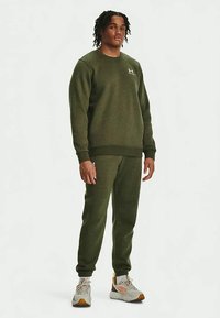 Νεαρός άνδρας που στέκεται φορώντας ελαιώδους πράσινου χρώματος φούτερ Under Armour και συνδυασμένα φόρμες με λευκά και πορτοκαλί αθλητικά παπούτσια σε απλό φόντο.