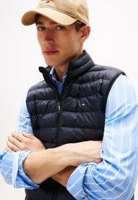 Colete acolchoado azul-marinho com gola alta e fecho de zip, usado sobre uma camisa azul-claro às riscas. Um boné castanho completa o conjunto.